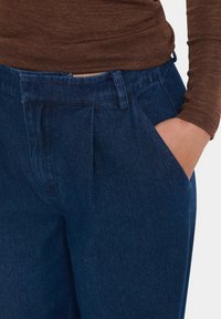 Tamsiai mėlynos džinsinės kelnės su raukšlėtu priekiu, šoninėmis kišenėmis ir klasikiniu juosmeniu. Audinys turi lygų tekstūrą su subtiliu blizgesiu.