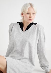 Sivý oversized sweatshirt šaty s výstrihom do V, pod ktorými je čierna košeľa s golierom. Mäkka tkanina a uvoľnený strih. Viditeľné strieborné náušnice.