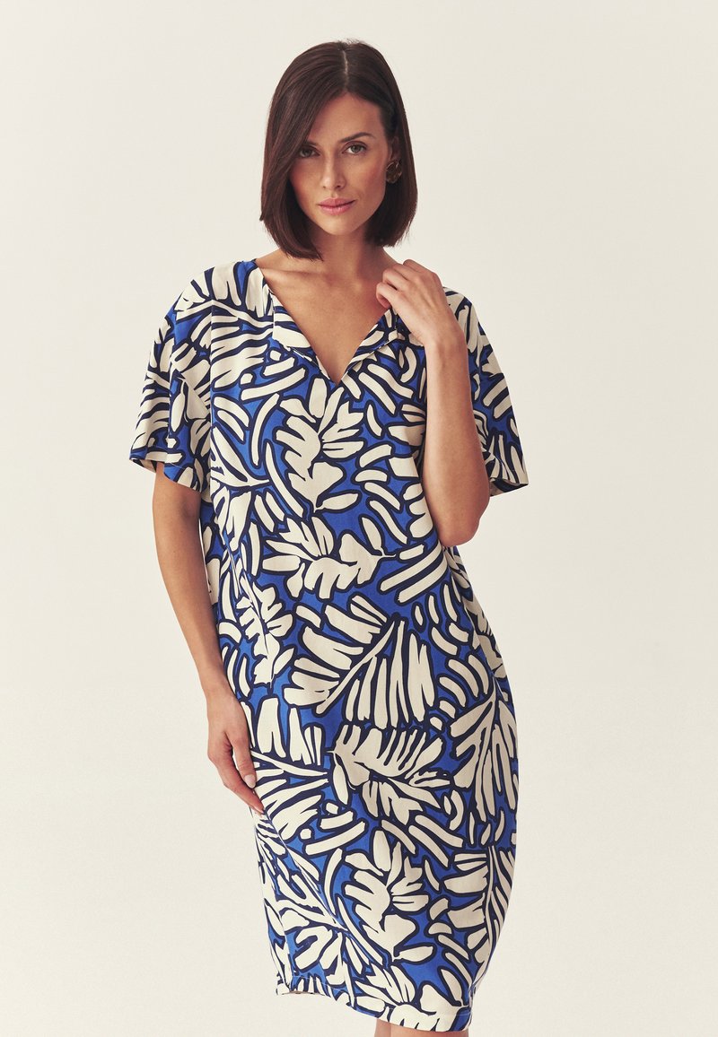 Femme aux cheveux courts et foncés portant une robe mi-longue à manches courtes avec un motif feuillu bleu et blanc, debout devant un fond uni.