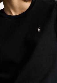 Černý bavlněný svetr s kulatým výstřihem, ribbovanou texturou a malým bílým vyšitým logem na hrudi. Jednoduchý design a střih.