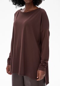 Bruine lange mouwen shirt van zachte stof, met een losse pasvorm, brede halslijn en gestikte zoom. Gecombineerd met slanke, donkergrijze textuurbroek.