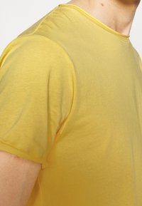 Gros plan d'une personne portant un t-shirt jaune uni à manches courtes, montrant les détails de l'épaule, du cou et du haut du bras.