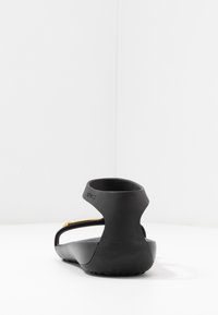 Musta sandaali, jossa on sileä kumimateriaali, avonainen takasuunnittelu, ohuita hihnoja ja pieni kultaisa yksityiskohta kannan lähellä. Tasainen pohja.