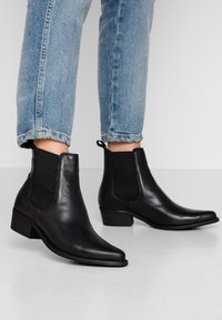 Bottines en cuir noir avec des panneaux latéraux élastiques, bout pointu et petit talon empilé, portées avec des jeans retroussés. Texture lisse.