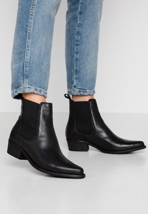 Bottines en cuir noir avec des panneaux latéraux élastiques, bout pointu et petit talon empilé, portées avec des jeans retroussés. Texture lisse.
