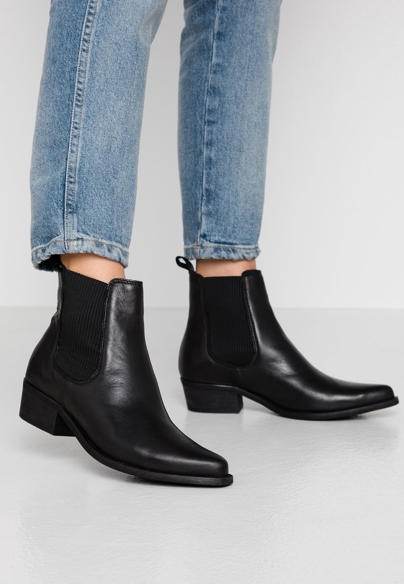 Bottines en cuir noir avec des panneaux latéraux élastiques, bout pointu et petit talon empilé, portées avec des jeans retroussés. Texture lisse.