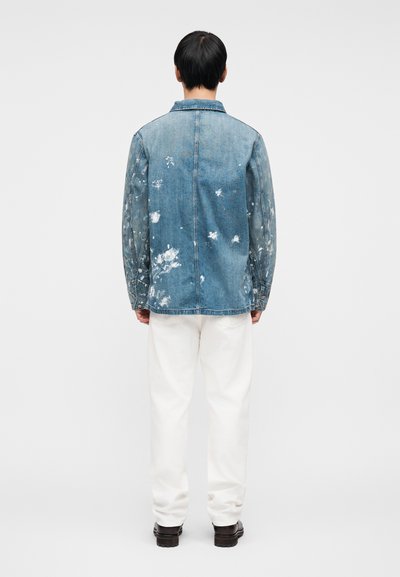 Τζιν μπουφάν με ανοιχτό μπλε ξέβαμμα, φθαρμένες λεπτομέρειες και λευκές πιτσιλιές βαφής στο πίσω μέρος. Συνδυάζεται με λευκό παντελόνι και μαύρα παπούτσια.