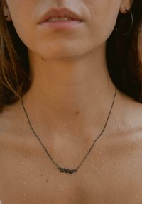 Dünne, dunkle Metallkette mit einem geschwungenen "Wellen"-Anhänger, getragen auf einem nackten Schlüsselbein. Die Haut hat ein nasses Aussehen mit sichtbaren Wassertropfen.