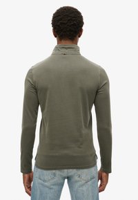 Camisa de manga larga en color verde oliva con cuello, que presenta aberturas laterales en el dobladillo y un pequeño logo en la parte posterior del cuello.
