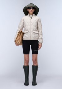 Beige gewatteerde jas met twee voorkruiszakken, zwarte fietsshorts, groene rubberen laarzen en een bijpassende hoed met brede rand. Draagt een beige tas.