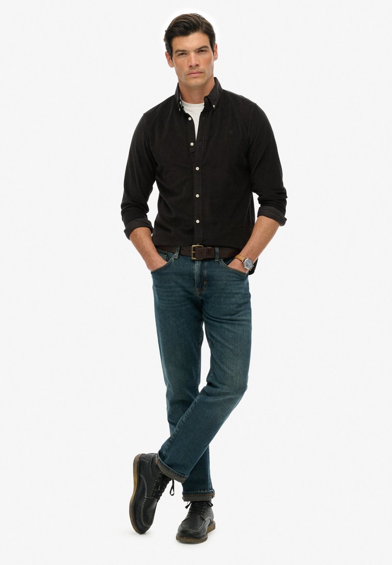 Mand stående med hænderne i lommerne iført sort skjorte med knapper, blå jeans, sorte sko og et armbåndsur mod hvid baggrund.
