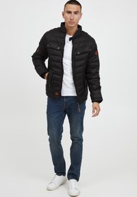 Black puffer jacket with a high collar, zip front, and two chest pockets. Paired with blue jeans and white sneakers.
