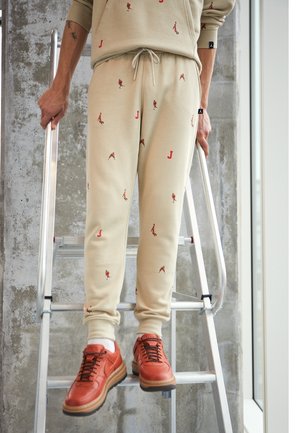 Person trägt beige Sweatpants mit roten Figuren und roten Sneakers, steht auf einer Metallleiter vor einer strukturierten Wand.