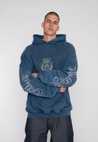 Hoodie surdimensionné bleu avec un graphique à l'avant et un script blanc sur les manches. Le tissu semble doux ; il dispose d'une capuche avec cordon de serrage et de poignets en rib genoemd.