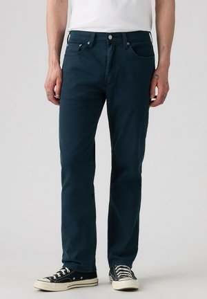 Mand iført mørkeblå jeans med lige ben, sorte og hvide sneakers og en hvid skjorte, står mod en ensfarvet baggrund.
