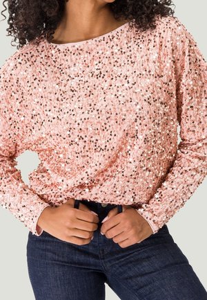 Langärmliges Oberteil aus hellrosa Stoff, das mit runden Pailletten bedeckt ist. Es hat einen weit ausgeschnittenen Hals und einen lässigen Schnitt und wird mit dunklen Jeans kombiniert.
