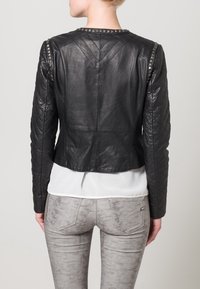 Giacca di pelle nera con texture trapuntata, dettagli in metallo argentato sulle spalle, vestibilità corta, abbinata a pantaloni grigi chiari a fantasia.