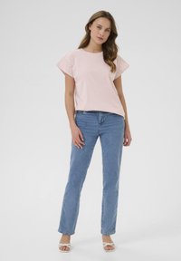 T-shirt rose clair à manches courtes avec une bordure en dentelle, associé à un jean droit bleu et des sandales à talons blancs.