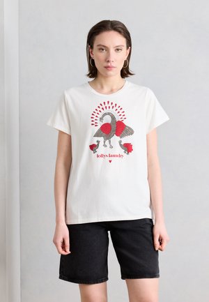 Fiatal nő fehér pólóban, amelyen piros-szürke madár motívum és a "lollys laundry" felirat látható, fekete rövidnadrággal, egyszerű fal előtt.