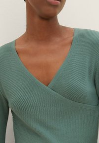 Jasnozielony prążkowany sweterek z głębokim dekoltem w kształcie litery V i kopertowym krojem. Charakteryzuje się miękką fakturą oraz bezszwowymi liniami przy dekolcie i ramionach.