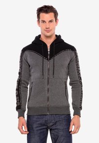 Graue und schwarze Kapuzenjacke mit Reißverschluss, ausgestattet mit einer Kapuze, einem Chevron-Design, metallischen Akzenten, seitlichen Taschen und bedrucktem Text auf den Ärmeln.