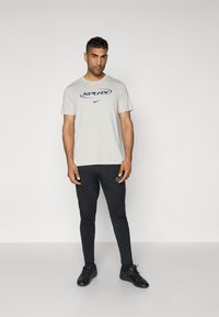 Lys grå t-shirt med "SPURS" print i marineblå, parret med sorte slim-fit bukser med et lille Nike-logo, og sorte træningssko.