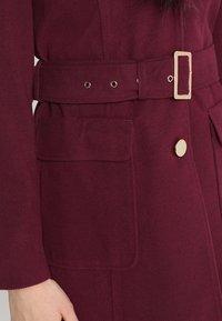 Manteau bordeaux en tissu texturé, avec une ceinture munie d'une boucle rectangulaire et une grande poche plaquée ornée d'un bouton rond.
