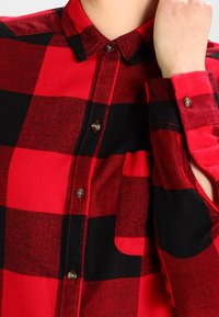 Camicia a quadri rossi e neri con colletto, taschino sul petto e maniche lunghe, indossata da una persona mentre sistema il polsino.