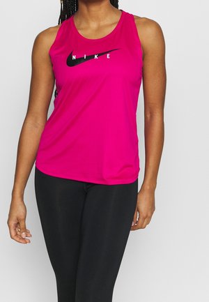 Donna che indossa un top Nike senza maniche di un rosa acceso e leggings neri su uno sfondo chiaro e uniforme.