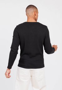 Camiseta negra de manga larga con un diseño texturizado y con relieve. Presenta un cuello redondo y un corte ligeramente ajustado. Camiseta blanca visible en el dobladillo.