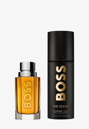 Le parfum Boss The Scent se présente dans un flacon rectangulaire en ambre avec un capuchon en métal argenté, accompagné d'un aérosol de déodorant noir mat.