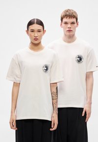 Kremno bele bombažne majice s kratkimi rokavi, s krožnim črno-belim logotipom na levi strani prsi, kombinirane s črnimi hlačami.