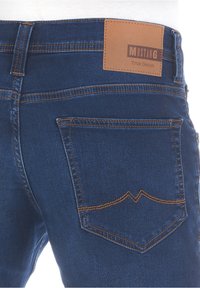 Jeans de mezclilla azul con una etiqueta de cuero que dice "Mustang True Denim", con costuras naranjas en contraste y un diseño en el bolsillo trasero.