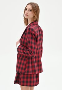 Blazer a quadri rossi e neri con un design strutturato, rever standard e una vestibilità rilassata, abbinato a una gonna a quadri coordinata.