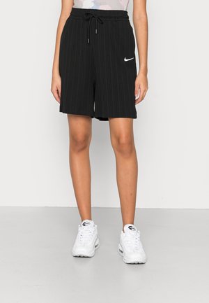 Personne portant un short Nike noir jusqu’aux genoux avec une taille à cordon et des baskets blanches Nike Air Max, debout devant un fond uni.