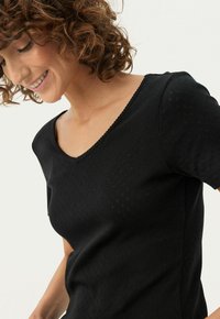 Sort kortærmet top med V-udskæring og struktureret mønster. Blødt materiale med en subtil glans, der fremhæver det minimalistiske design.