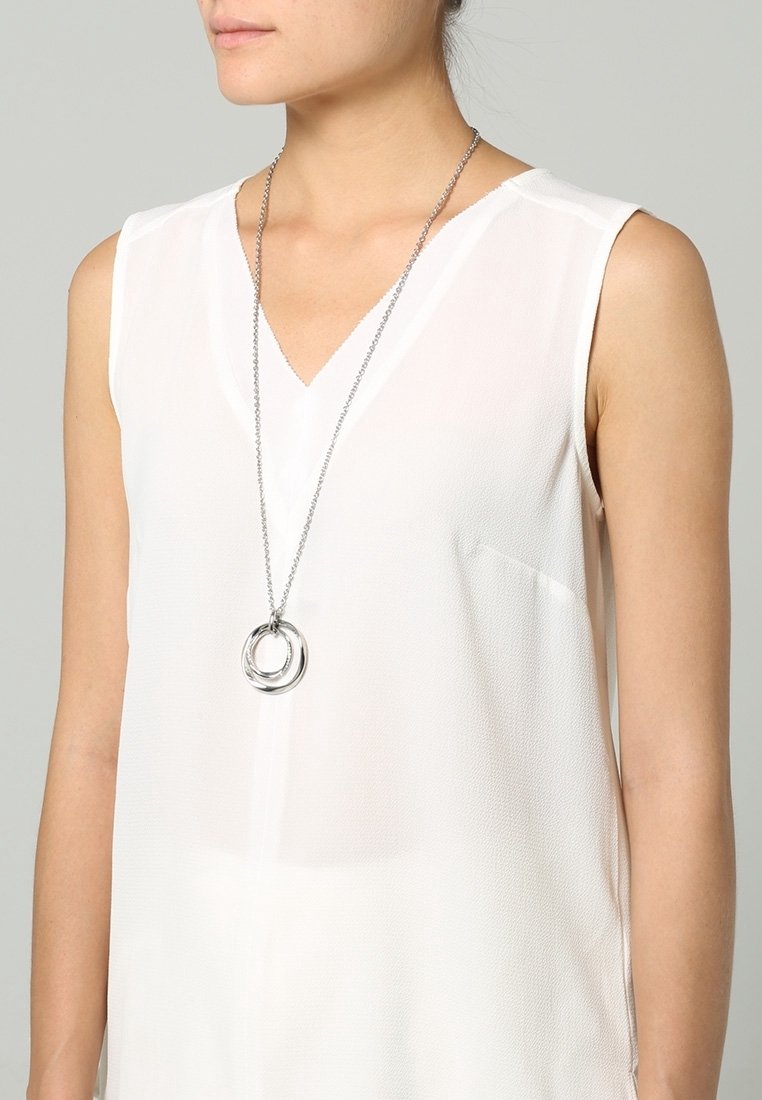 Femme portant un haut blanc sans manches avec un col en V et un long collier en argent tenant deux anneaux entrelacés en pendentif.