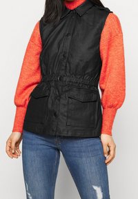 Musta hihaton liivi, jossa on napillinen etuosa, sä�ädettävä vyötärö ja kaksi etutaskua, pidetty kirkkaanpunaisen neuleen päällä. Alla näkyvät farkut.