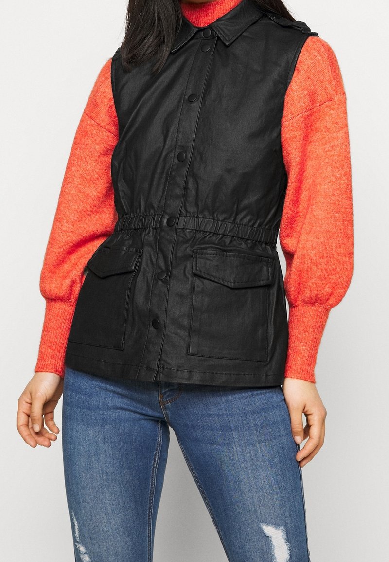 Musta hihaton liivi, jossa on napillinen etuosa, säädettävä vyötärö ja kaksi etutaskua, pidetty kirkkaanpunaisen neuleen päällä. Alla näkyvät farkut.