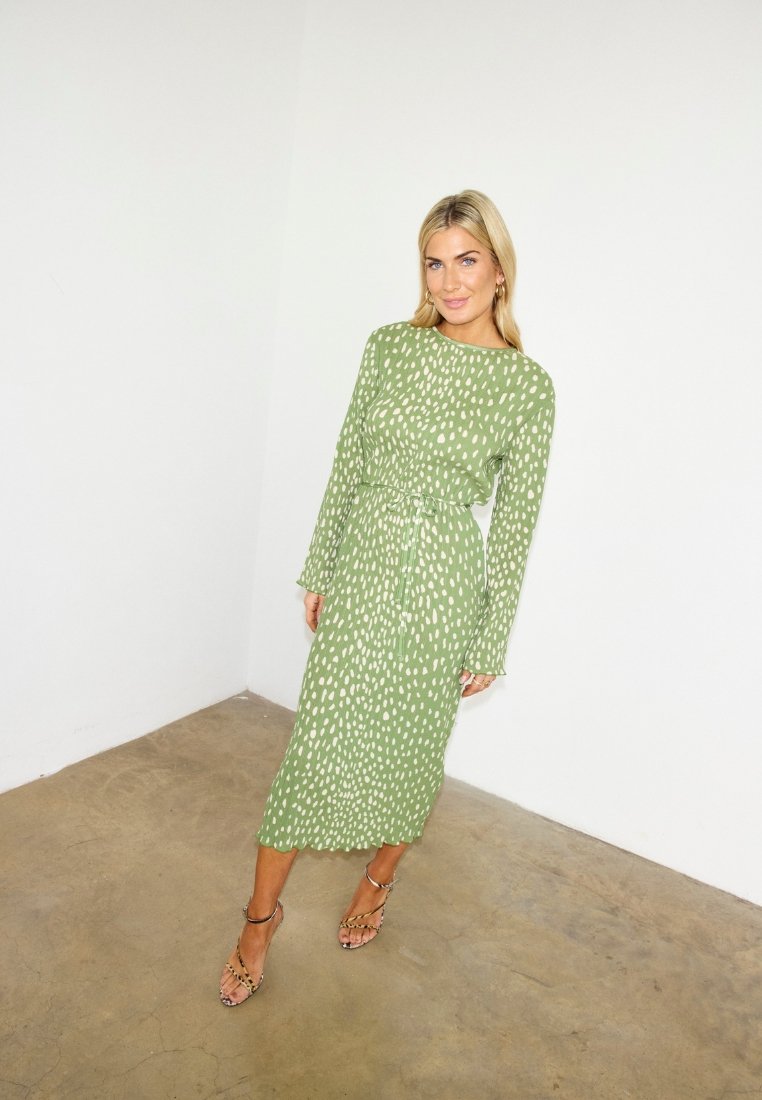 Γυναίκα με μακρυμάνικο πράσινο φόρεμα με λευκές βούλες μέχρι το μέσο της γάμπας και ψηλοτάκουνα παπούτσια με λουράκι στον αστράγαλο, στέκεται μπροστά σε λευκό τοίχο.