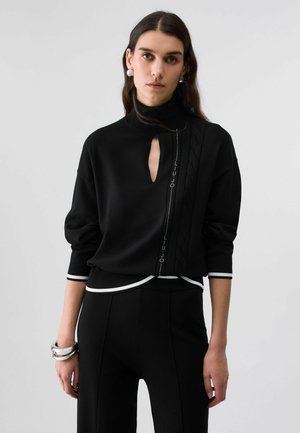 Vrouw met lang donker haar draagt een zwarte trui met een keyhole-halslijn en decoratieve verticale streep, gecombineerd met zwarte broek en zilveren oorbellen.