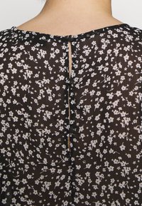 Blouse noire à motifs floraux blancs, tissu translucide, col rond et fermeture à boutons dans le dos avec une petite fente.