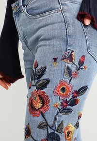 Jeans de mezclilla en azul claro con bordado floral en rojo, amarillo y negro. Presenta un diseño ajustado y un estilo clásico de cinco bolsillos.