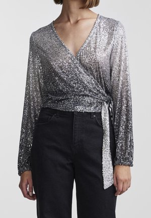 Vrouw draagt een zilverkleurige pailletten lange mouwen wikkeltop met V-hals, gecombineerd met zwarte broek met hoge taille op effen achtergrond.