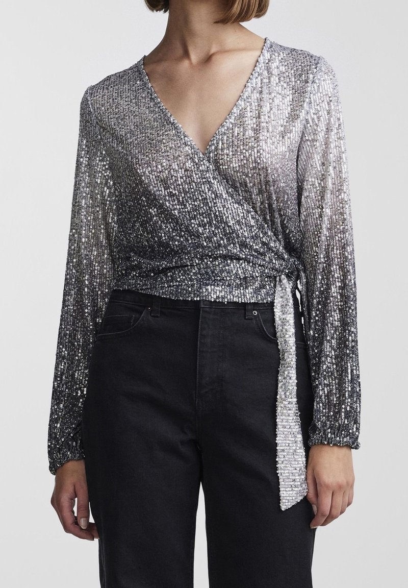 Femme portant un haut cache-cœur à manches longues en sequins argentés avec décolleté en V, associé à un pantalon noir taille haute sur fond uni.