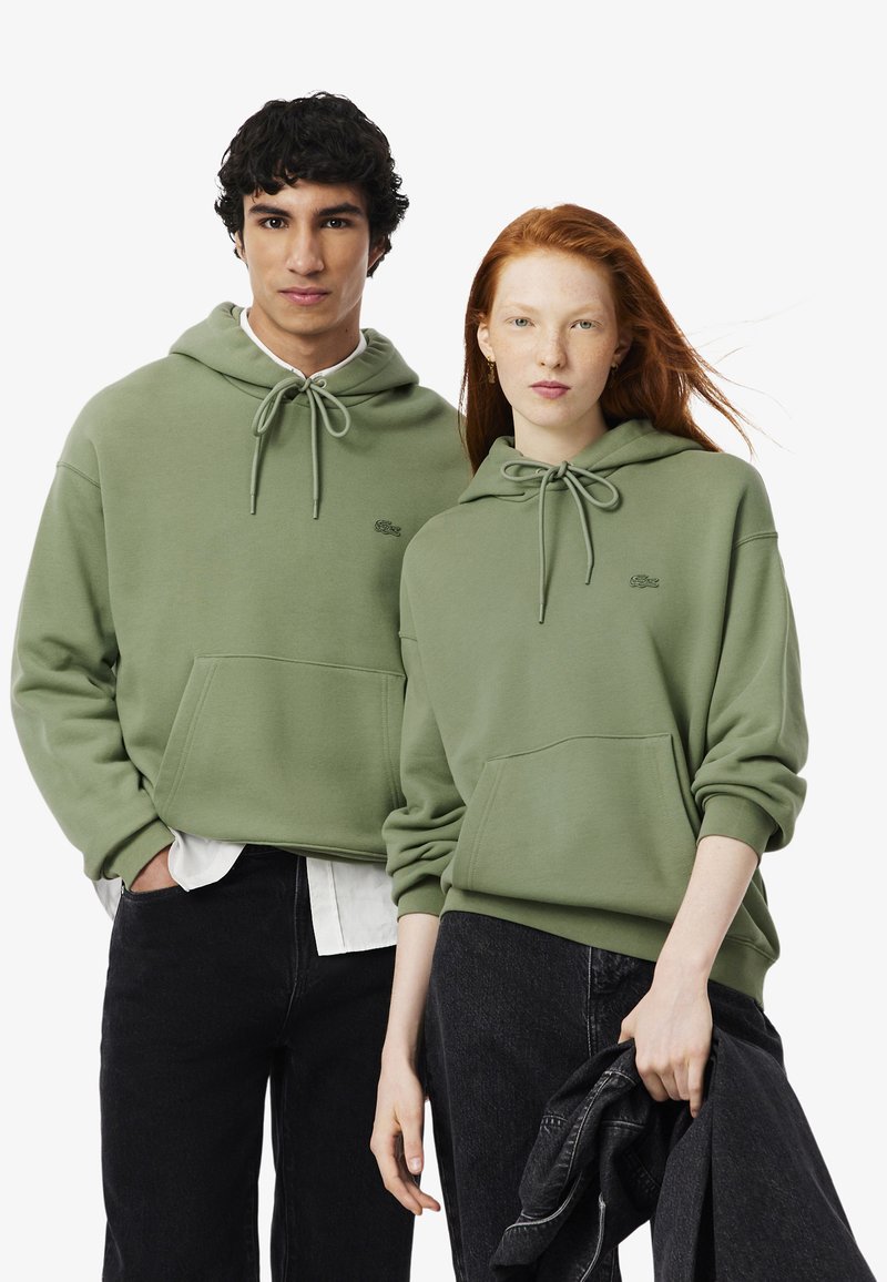 Felpe verde oliva con cappuccio regolabile, tasche a marsupio e logo ricamato. Indossate da due persone che portano jeans neri.
