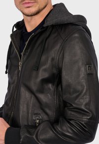 Sort læderjakke med grå hætte, lynlås foran, ribbede manchetter og et accentpatch på ærmet. Glat tekstur, tætsiddende design.