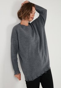 Szary, oversize sweter z prążkowanym dekoldem i postrzępionym dołem. Materiał wydaje się miękki, z luźnymi rękawami i subtelną teksturą.