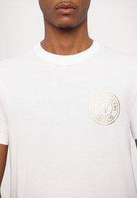 Valge puuvillane T-särk ümmarguse kaelusega. Vasakul rinnal on kuldne embosseeritud ringikujuline logo, millel on tekst "Versace Jeans Couture."