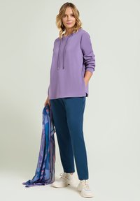 Felpa con cappuccio viola dotata di lacci, abbinata a pantaloni jogger blu. Il modello tiene in mano una sciarpa multicolore. Sneakers bianche completano il look.